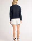 A woman is shown from behind wearing a dark hand- crochet sweater, white shorts, and beige wedge sandals against a plain light gray studio background.