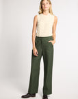 A woman modeling a sleeveless cream cable-knit top and dark olive green wide-leg trousers, paired with dark brown leather shoes, against a light gray studio background.