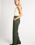A woman modeling a pair of high-waisted, wide-leg olive green trousers, paired with a cream cable-knit sleeveless top and dark brown leather loafers, against a plain light gray studio background.