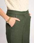 A close-up of a person wearing dark olive green wide-leg trousers with a cream cable-knit top. The person's hand is in a pocket, wearing a chunky gold chain bracelet, against a light gray background.
