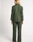 A woman shown from the back wearing a matching olive green suit, featuring a structured blazer with button details on the cuffs and wide-leg trousers, set against a plain light gray studio background.