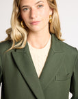 A woman with blonde hair wearing a dark olive green blazer over a cream cable-knit sweater, accessorized with gold hoop earrings, against a plain light gray background.