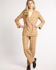 A woman modeling a beige and white windowpane check suit, consisting of a double-breasted blazer and matching trousers, paired with black leather shoes, against a light gray studio background.