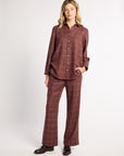 A woman modeling a matching two-piece plaid lounge set in a dusty rose-brown color with thin tan windowpane lines, consisting of a long-sleeve button-up shirt and wide-leg pants, paired with black leather shoes against a light gray studio background.