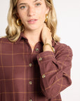 A woman wearing a brown and rust plaid button-down shirt with gold buttons, styled with gold hoop earrings and a gold bracelet against a plain white background.