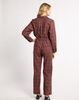 A woman is shown from the back wearing a matching long-sleeve collared jumpsuit in a dark reddish-brown color with a thin windowpane plaid pattern. The jumpsuit has an elastic waist and straight legs, paired with dark brown shoes, against a plain light gray studio background.