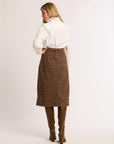 Back of a woman with hand on hip wearing a white skirt and brown plaid skirt with brown boots against a white background