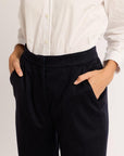 Close up of woman wearing a white shirt and navy pants on a plain background with hands in pockets