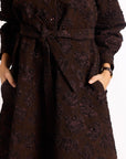 Close up of brown brocade dress with tie belt and hands in pockets