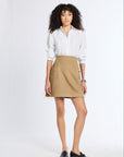 A woman with curly dark hair stands against a white studio background wearing a white button-up shirt, a tan mini skirt, and black loafers.