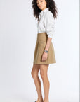 A woman modeling a white long-sleeve button-up shirt and a beige A-line mini skirt, paired with black loafers, against a plain white studio background.