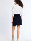 A woman is shown from behind wearing a white long-sleeve button-up shirt and a dark navy blue corduroy mini skirt, paired with black flat shoes, against a plain white studio background.