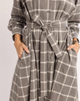 Woman with hands in pockets wearing flannel gray and white checkered dress with a tie waist on a plain background