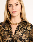 Woman wearing a black and gold floral patterned blouse with a gold necklace against a plain background