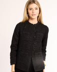 Woman wearing a black textured jacket against a plain background wearing gold hoops