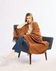 Woman sitting on a chair wearing a brown blanket, blue jeans, and white shoes against a white background