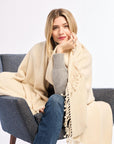 Woman sitting on a gray chair wearing a beige blanket and gray sweater against a white background