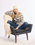 Woman sitting on a gray chair with a beige blanket, wearing a patterned sweater and jeans.