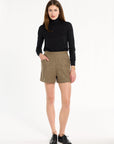 A woman modeling the Painter's Fleck Herringbone Archer Shorts with a black turtleneck and black loafers, front view, photographed against a plain white studio background.