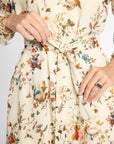 Close-up of a person wearing a cream-colored long-sleeve dress with a multicolor floral and bird print, featuring a tie waist belt and pockets.