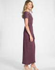 A woman modeling a short-sleeved, floor-length purple dress with a tie waist, shown in profile against a light gray studio background.