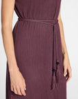 A close-up of a woman wearing a ribbed purple dress with a matching tie belt and tassels, shown against a plain light background.