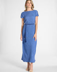 A woman modeling a blue pleated dress with a tassel around the waist on a white backdrop