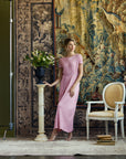 A woman models a light pink midi dress with short sleeves and a gathered waist, standing in a styled studio setting with a large tapestry backdrop, a pedestal with flowers, and a white armchair.