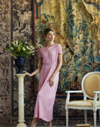 A woman models a light pink midi dress with short sleeves and a gathered waist, standing in a styled studio setting with a large tapestry backdrop, a pedestal with flowers, and a white armchair.