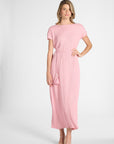 A woman modeling a light pink dress with short sleeves, a matching drawstring waist, and tassel details, set against a plain light gray studio background.