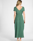 A woman viewed from the back wearing a green short-sleeve maxi dress with a V-neckline and tie waist, paired with beige heels, against a light gray studio background.