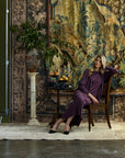 Woman sitting against rugs wearing a pleated purple silk cardigan with gemstone buttons