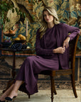 Woman sitting in a wooden chair against a patterned background wearing a pleated cardigan, skirt, and shell