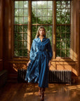 Woman in a blue dress standing in a room with large windows and wooden floors.