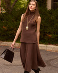 Woman in brown ribbed knit skirt and matching top holding a handbag on a street with bushes in the background