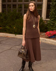 Woman in a brown ribbed knit sleeveless top and matching skirt standing outdoors with a building and flowers in the background