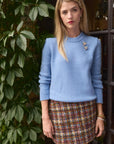 Woman wearing a light blue sweater and plaid tweed skirt standing next to a door with greenery.