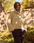 A woman wearing a cream-colored cowl-neck knit sweater and dark navy trousers, standing outdoors in a sun-dappled wooded setting.