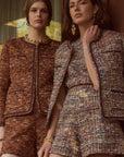 Two women modeling matching tweed mini suits in an outdoor setting with warm-toned glass doors and trees visible in the background. The suits feature a textured multicolor tweed pattern with brown, beige, and gold tones.