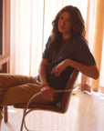 Woman in a house sitting in a chair modeling a blue cotton tee with brown pants