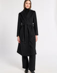 A woman modeling a long black wrap-style cashmere coat with a matching waist tie, worn over a black turtleneck and black wide-leg trousers, against a plain light gray studio background.