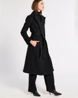 A woman modeling a long black belted coat, black wide-leg trousers, and black pointed-toe heels against a plain light gray studio background.