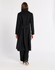 A woman viewed from behind wearing a long black belted coat, black pants, and black high-heeled shoes against a plain white studio background.