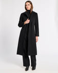 A woman modeling a long black coat over a black turtleneck and wide-leg black trousers, standing against a plain light gray studio background.