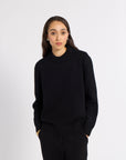A woman modeling a black crewneck knit sweater and matching black trousers against a plain light gray studio background.