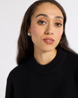 A close-up portrait of a woman with dark hair wearing a black ribbed knit mock neck sweater against a plain light gray background.