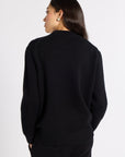 A woman shown from the back wearing a black knit sweater and black pants against a plain light gray studio background.