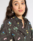 A woman wearing a black button-up shirt with a colorful floral and bee pattern, paired with white hoop earrings, photographed against a light gray background.