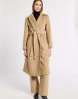 A woman modeling a long camel-colored cashmere coat with a matching belt and wide-leg trousers, styled with a black turtleneck and black pointed-toe heels against a plain white studio background.