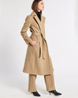 A woman modeling a long camel-colored belted coat with matching wide-leg trousers and black pointed-toe heels, photographed against a plain light gray studio background.